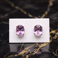 A pair of oval purple gemstone stud earrings in a gold setting, displayed on a small white card over a black marble surface with gold veining. Amethyst

