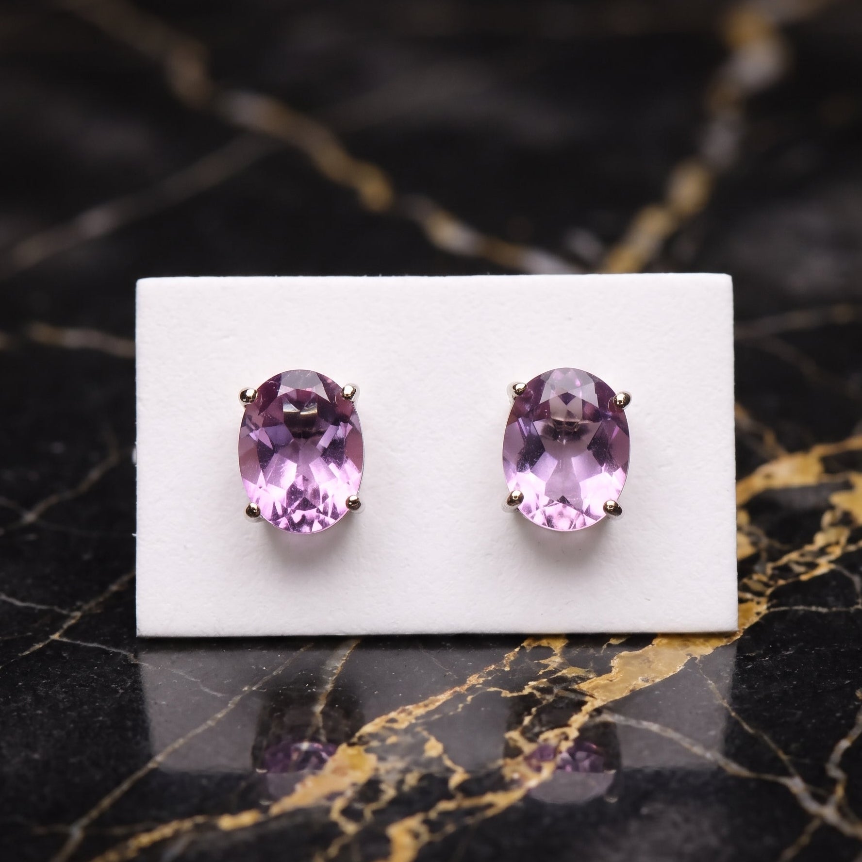 A pair of oval purple gemstone stud earrings in a gold setting, displayed on a small white card over a black marble surface with gold veining. Amethyst
