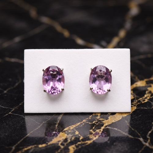 A pair of oval purple gemstone stud earrings in a gold setting, displayed on a small white card over a black marble surface with gold veining. Amethyst
