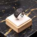 A silver-toned ring with a wide, heavily paved black band sits on a small wooden display block with a white cushion, placed on a black marble surface with gold veining.
