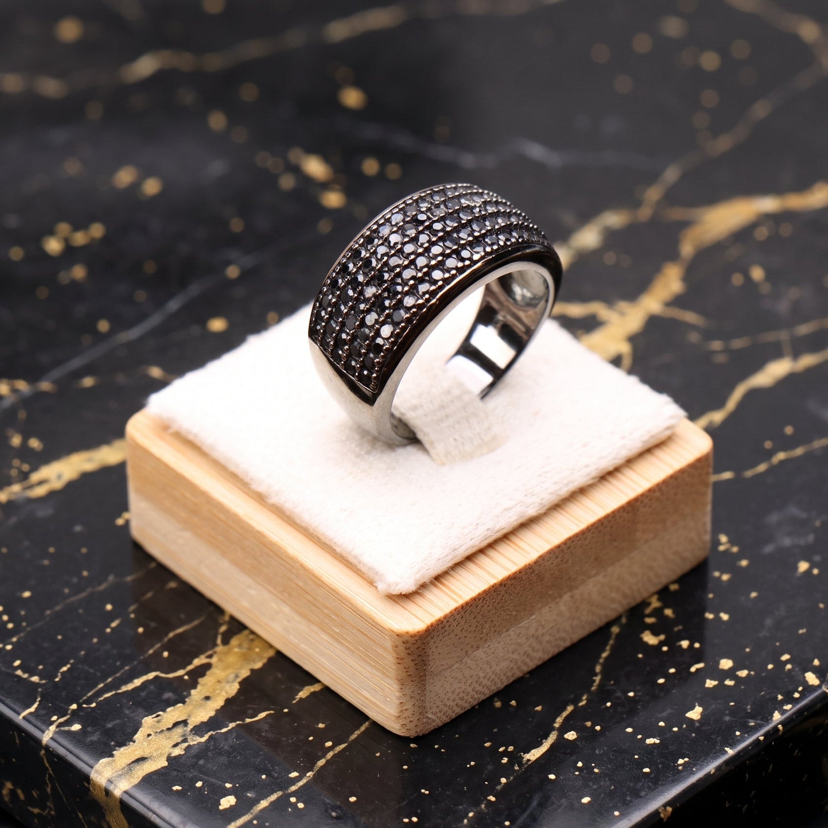 A silver-toned ring with a wide, heavily paved black band sits on a small wooden display block with a white cushion, placed on a black marble surface with gold veining.