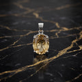An oval-cut yellow gemstone pendant in a silver-toned setting, displayed on a polished black marble surface with gold veining.