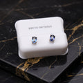 A pair of oval stud earrings with light blue stones in silver settings, displayed on a white jewelry box labeled 'STERLING SILVER' on a black marble surface with gold veining. tanzanite dainty

