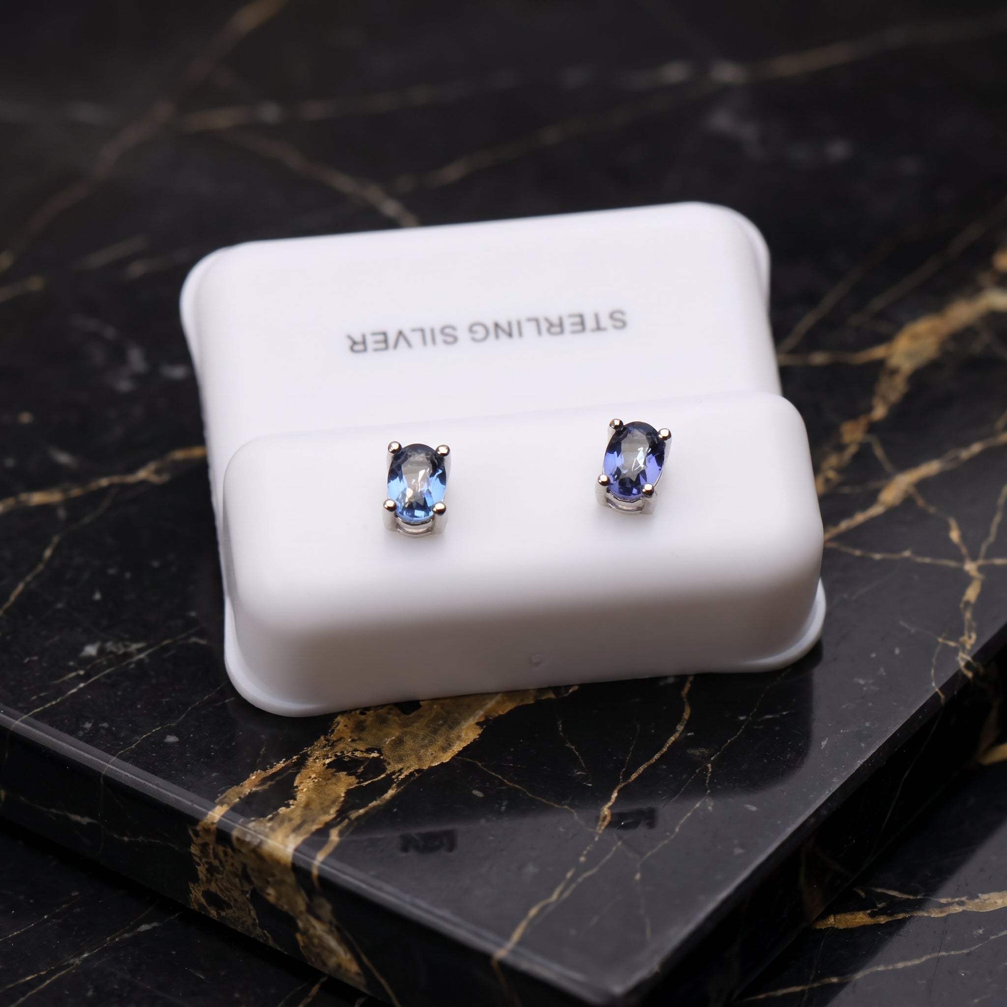 A pair of oval stud earrings with light blue stones in silver settings, displayed on a white jewelry box labeled 'STERLING SILVER' on a black marble surface with gold veining. tanzanite dainty
