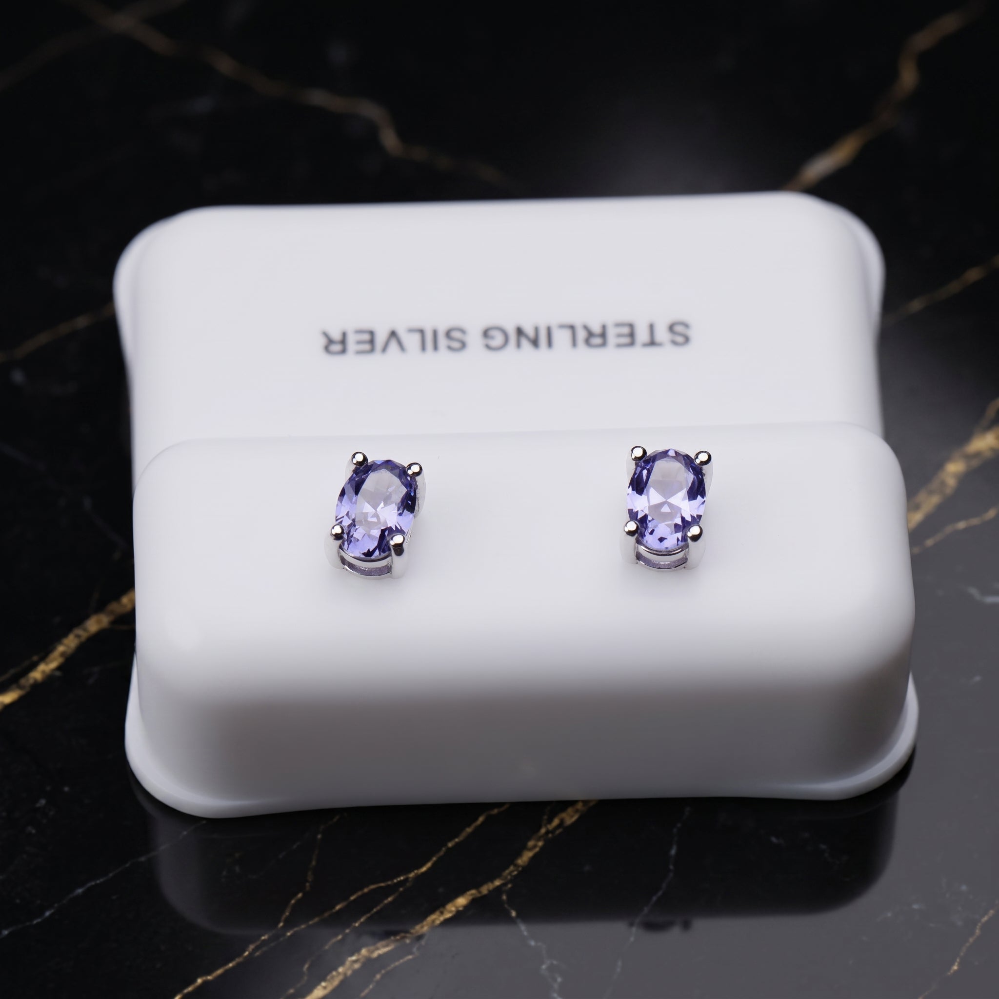 A pair of oval stud earrings with light purple gemstones in silver settings, displayed on a white jewelry box labeled 'STERLING SILVER' on a black marble surface. tanzanite