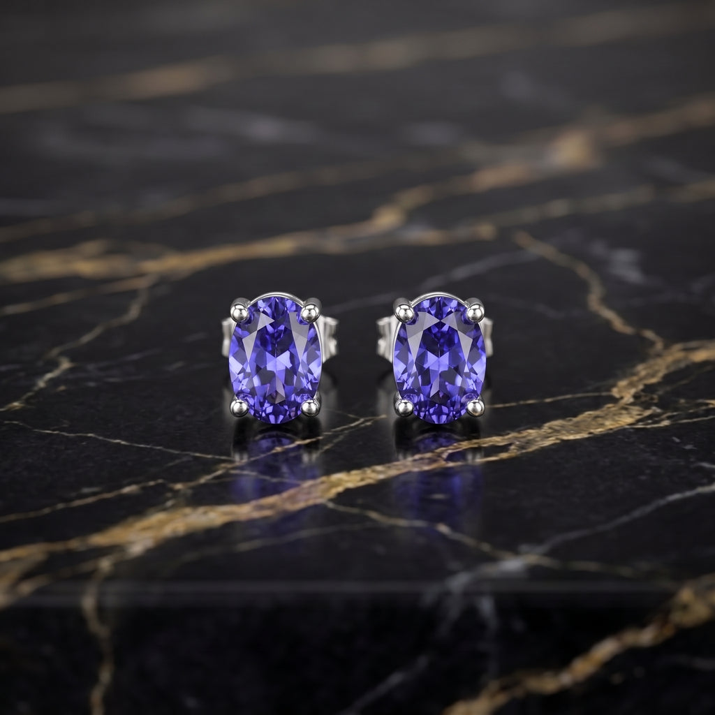 A pair of oval-cut tanzanite stud earrings in silver prong settings, displayed on a polished black marble surface with gold veining.