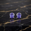 A pair of oval-cut tanzanite stud earrings in silver prong settings, displayed on a polished black marble surface with gold veining.