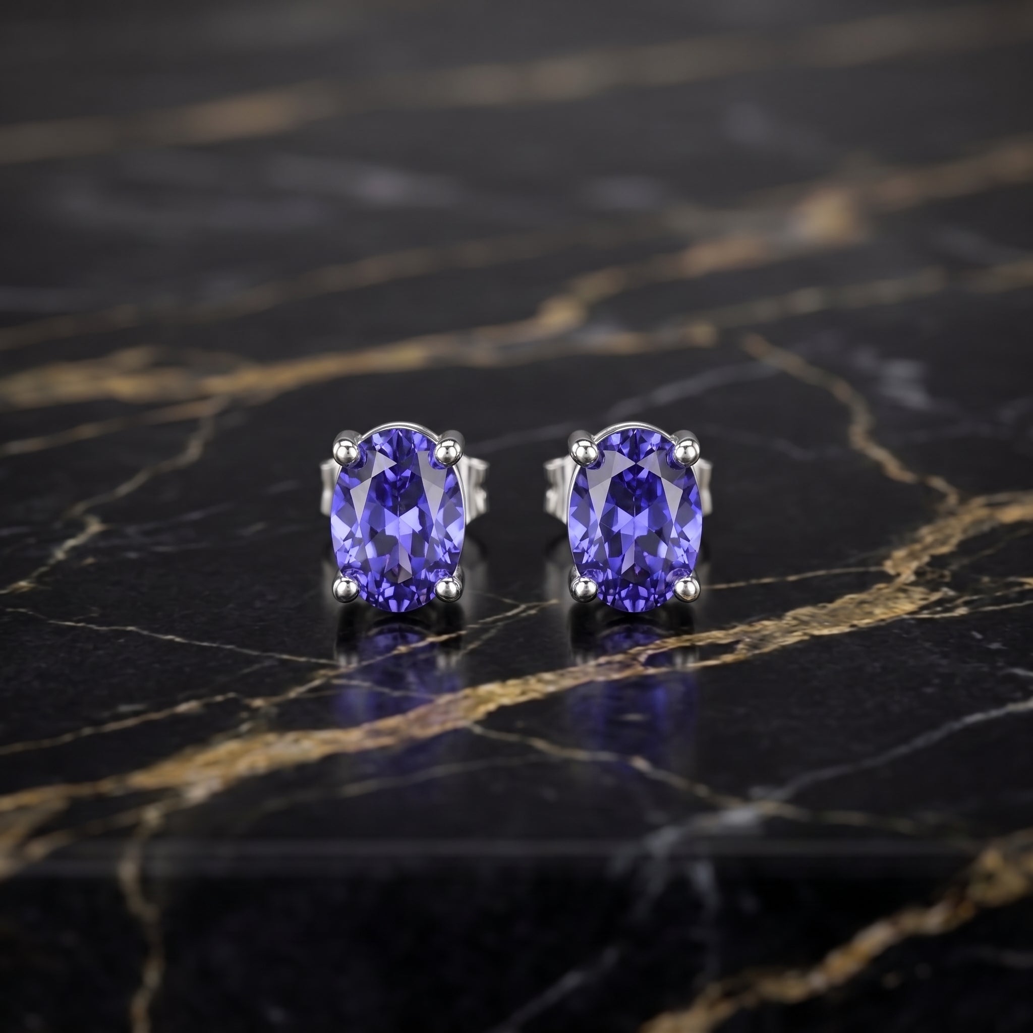 A pair of oval-cut tanzanite stud earrings in silver prong settings, displayed on a polished black marble surface with gold veining.