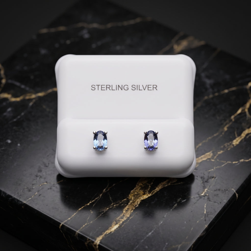 A pair of oval-cut light blue gemstone stud earrings in silver settings, presented on a white display card labeled 'STERLING SILVER' which rests on a black marble surface with gold veining.