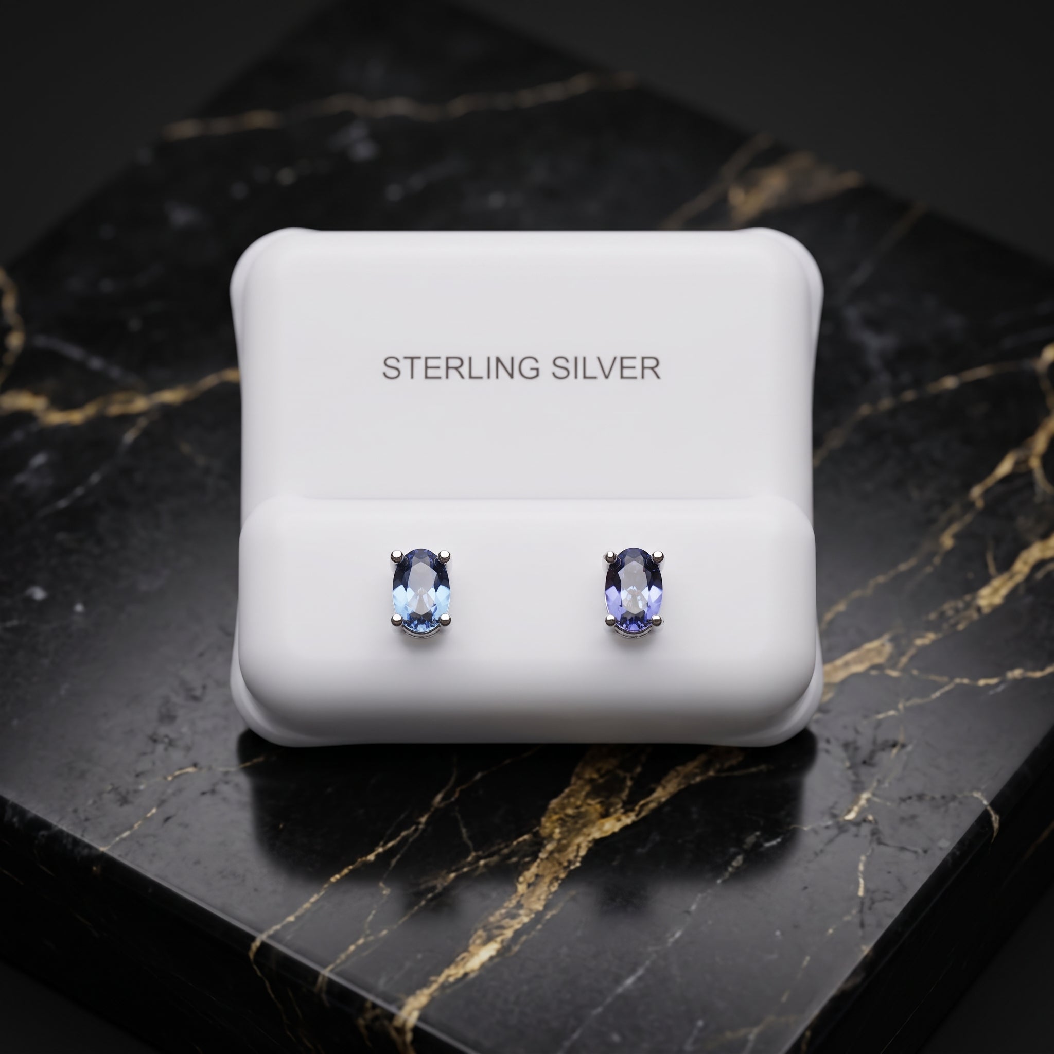 A pair of oval-cut light blue gemstone stud earrings in silver settings, presented on a white display card labeled 'STERLING SILVER' which rests on a black marble surface with gold veining.