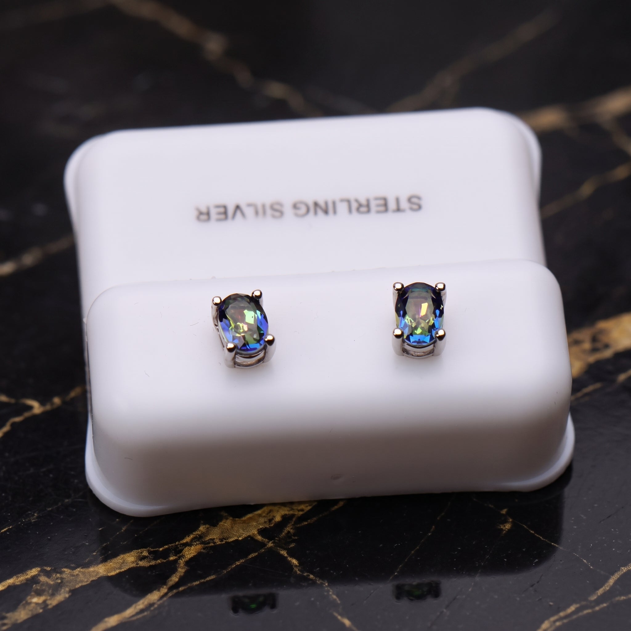 A pair of oval stud earrings with iridescent blue and green stones in silver settings, displayed on a white 'STERLING SILVER' box on a black marble surface.