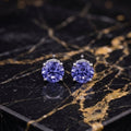 A pair of stud earrings with round, faceted purple-blue gemstones in silver settings, displayed on a black and gold veined marble slab. tanzanite
