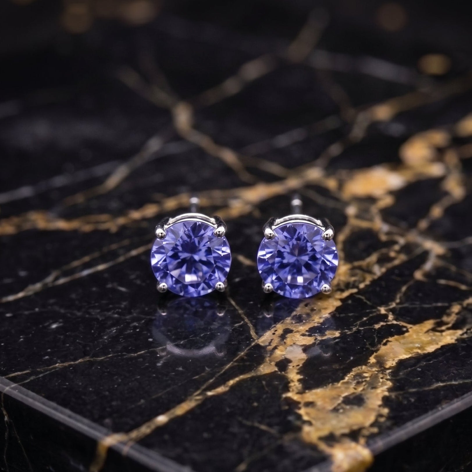 A pair of stud earrings with round, faceted purple-blue gemstones in silver settings, displayed on a black and gold veined marble slab. tanzanite

