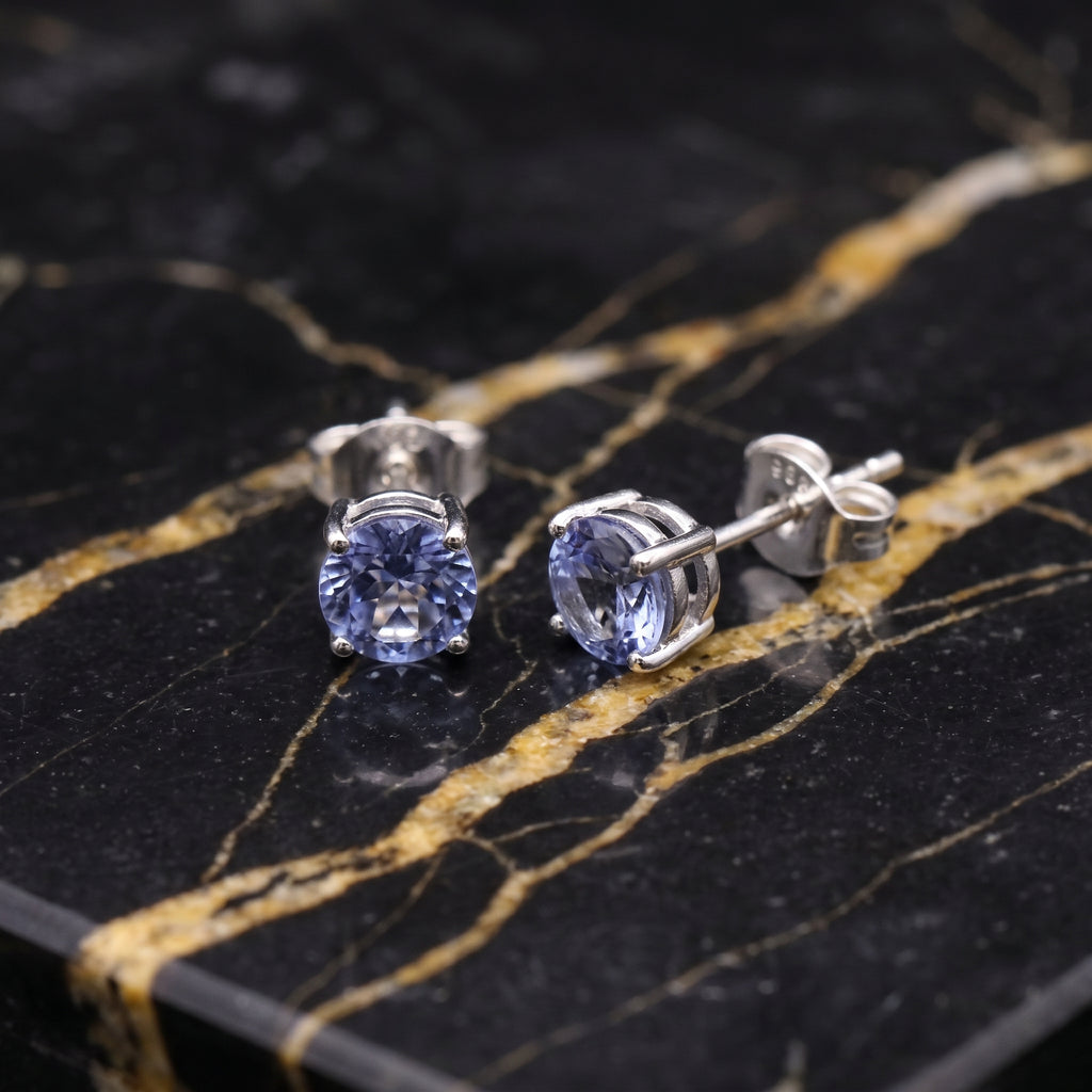 A pair of round-cut blue gemstone stud earrings in silver settings, displayed on a black and gold veined marble surface. tanzanite
