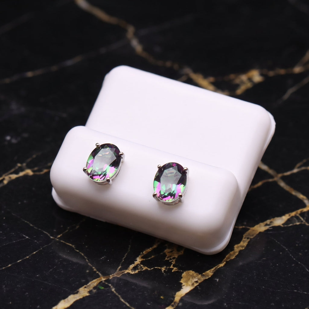 A pair of oval stud earrings with iridescent, rainbow-flecked clear gemstones in silver settings, displayed on a small white rectangular stand over a black marble surface with gold veining.