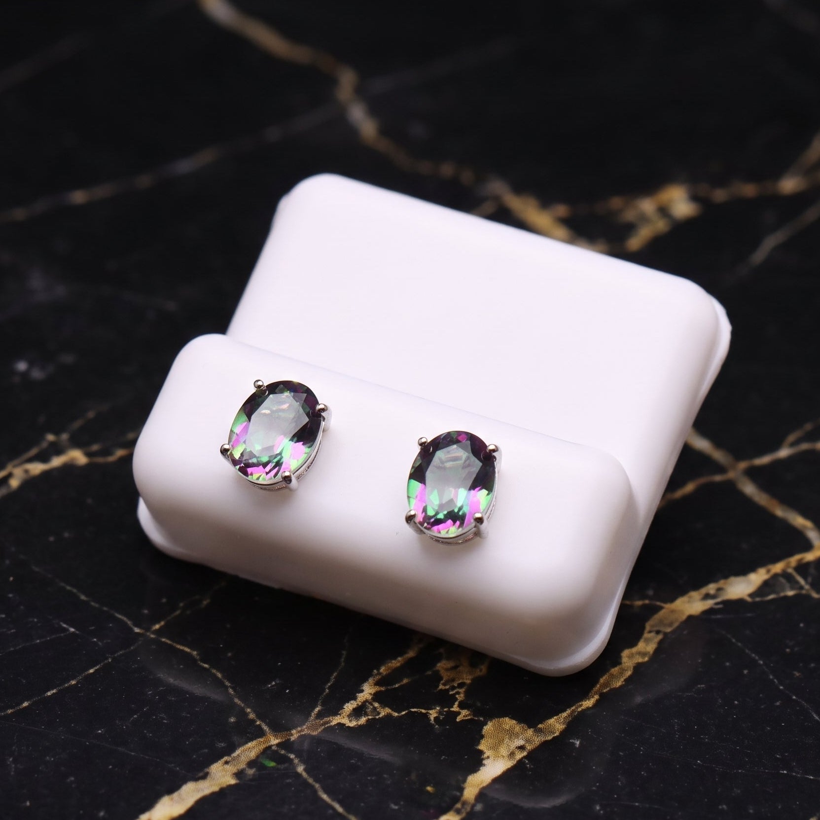 A pair of oval stud earrings with iridescent, rainbow-flecked clear gemstones in silver settings, displayed on a small white rectangular stand over a black marble surface with gold veining.