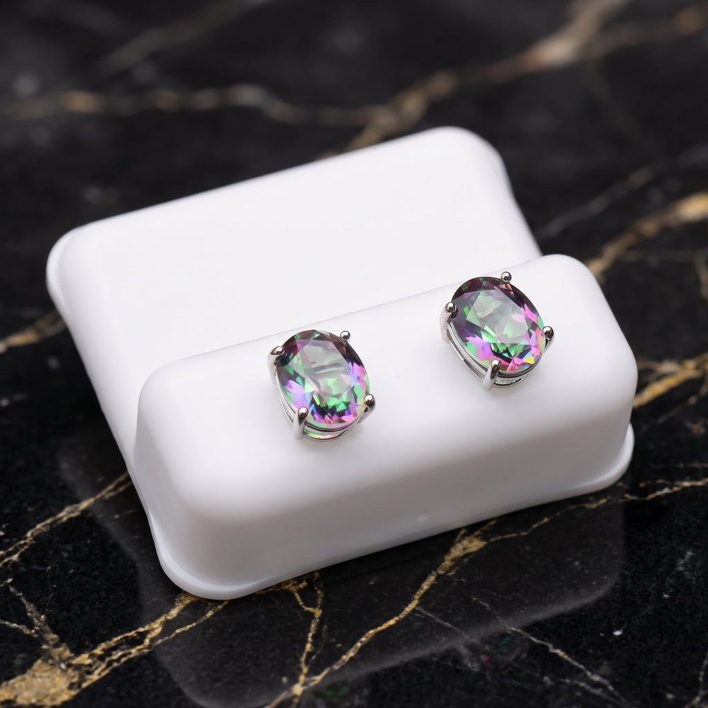 A pair of oval stud earrings with iridescent rainbow-colored gemstones in silver settings, displayed on a white rectangular stand over a black marble surface. Mystic Topaz