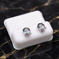 A pair of oval stud earrings with iridescent rainbow-colored gemstones in silver settings, displayed on a white rectangular stand over a black marble surface. Mystic Topaz