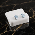 A pair of oval-cut light blue gemstone stud earrings in silver settings, displayed on a small white rectangular stand over a black marble surface with gold veining. Blue Topaz