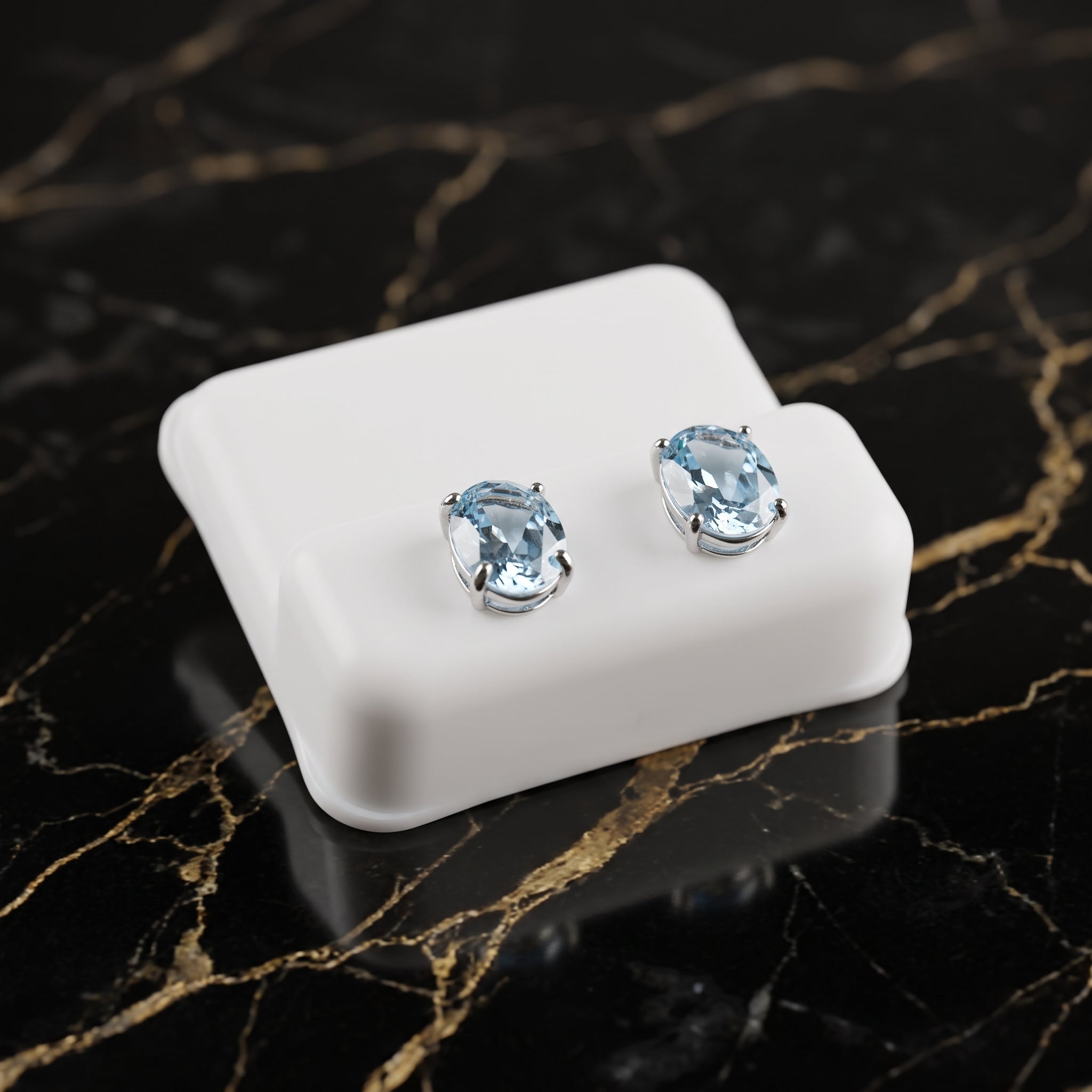 A pair of oval-cut light blue gemstone stud earrings in silver settings, displayed on a small white rectangular stand over a black marble surface with gold veining. Blue Topaz