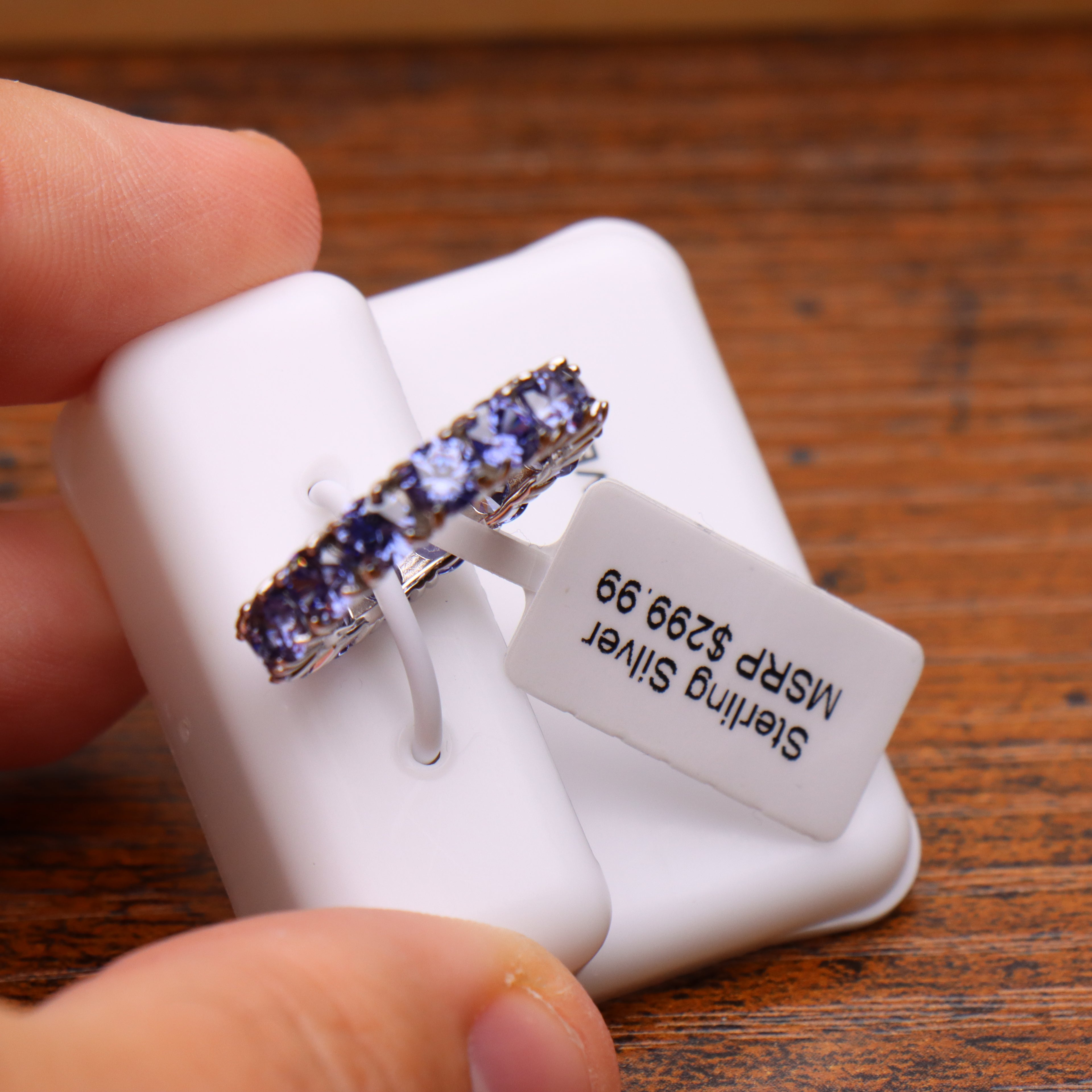 A sterling silver ring with a row of round blue gemstones is displayed on a white plastic ring holder, held by fingers over a wooden surface. The tag on the holder reads 'Sterling Silver MSRP $289.