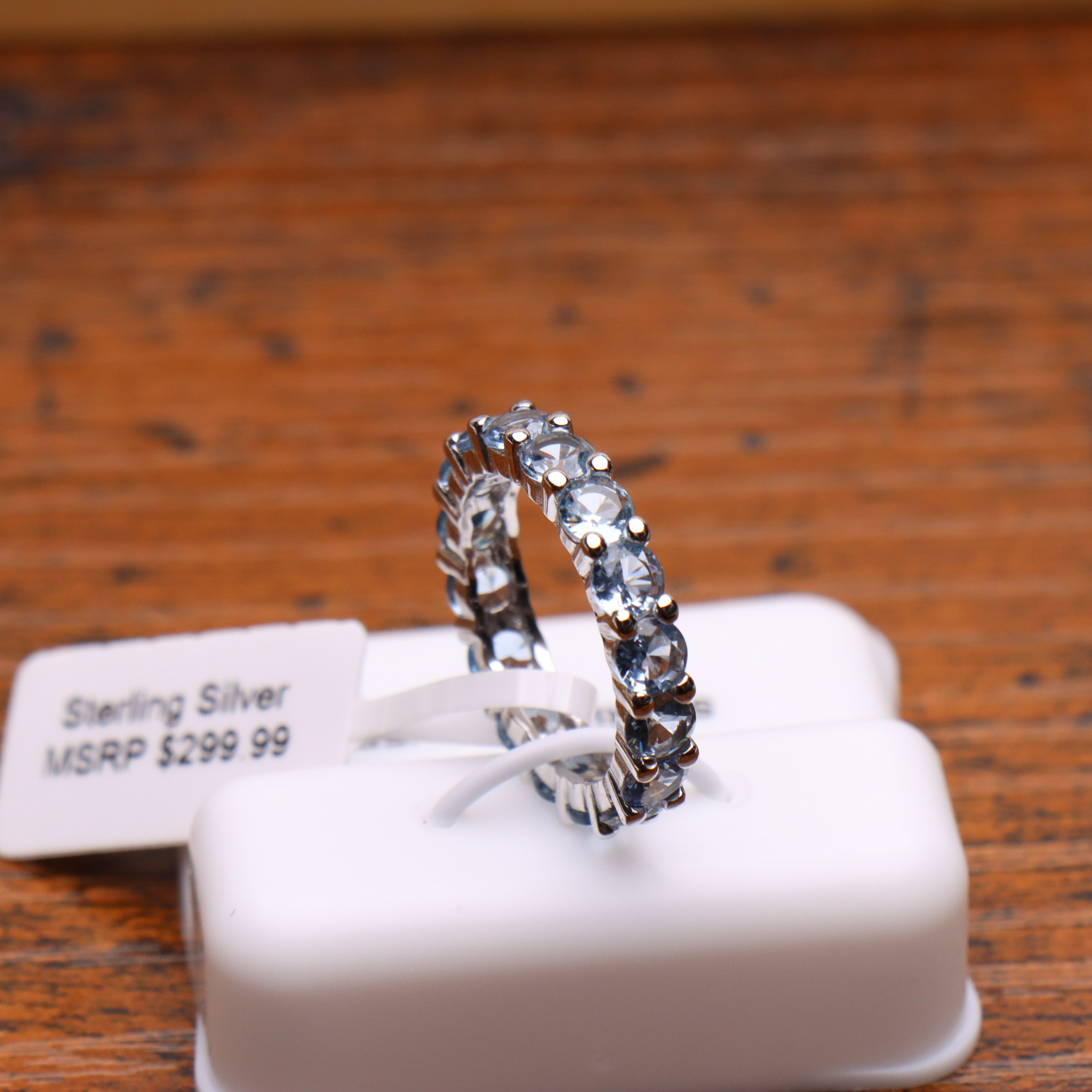 A sterling silver eternity ring with clear round-cut stones, displayed on a white plastic jewelry display block with a price tag that reads 'Sterling Silver MSRP $299.99' on a wooden table.