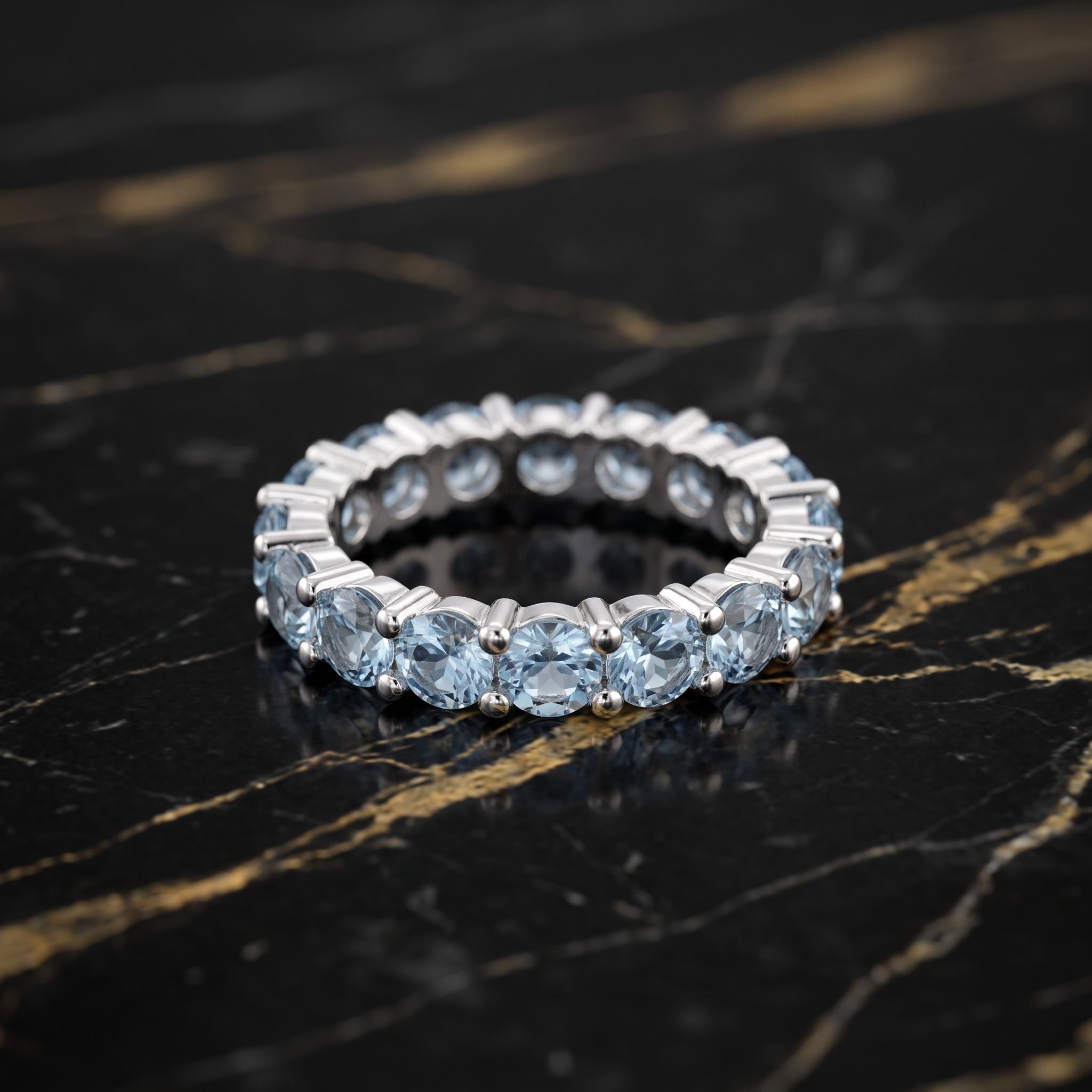 A silver eternity ring set with round light blue gemstones, resting on a black marble surface with gold veining.