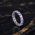 A silver eternity ring set with round-cut light purple amethyst gemstones, displayed on a black and gold veined marble surface.
