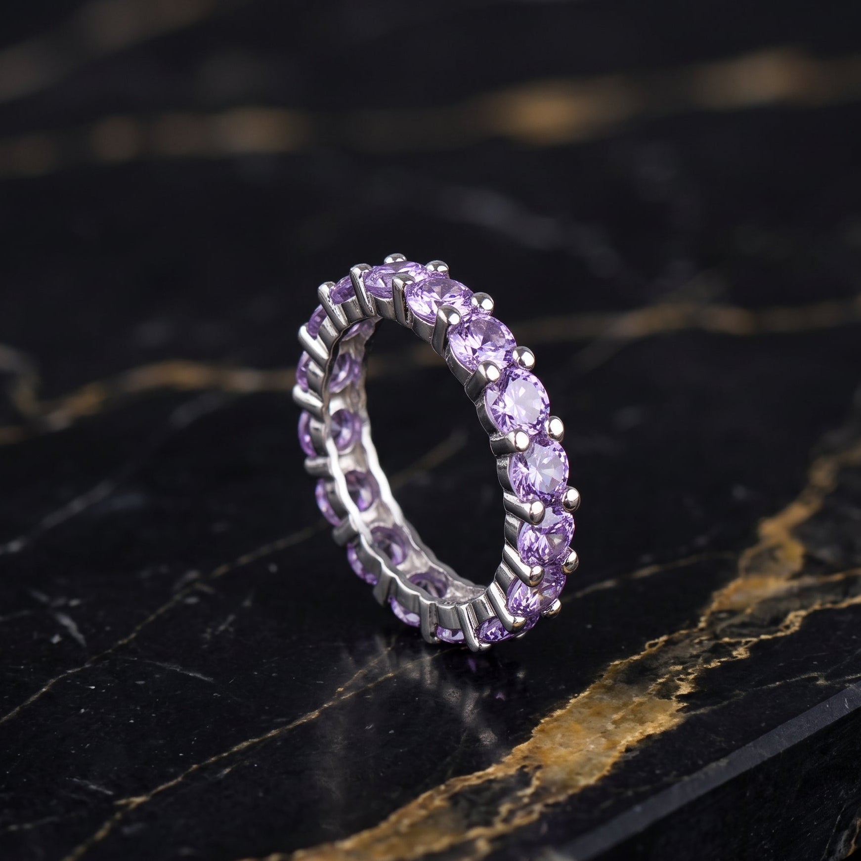 A silver eternity ring set with round-cut light purple amethyst gemstones, displayed on a black and gold veined marble surface.