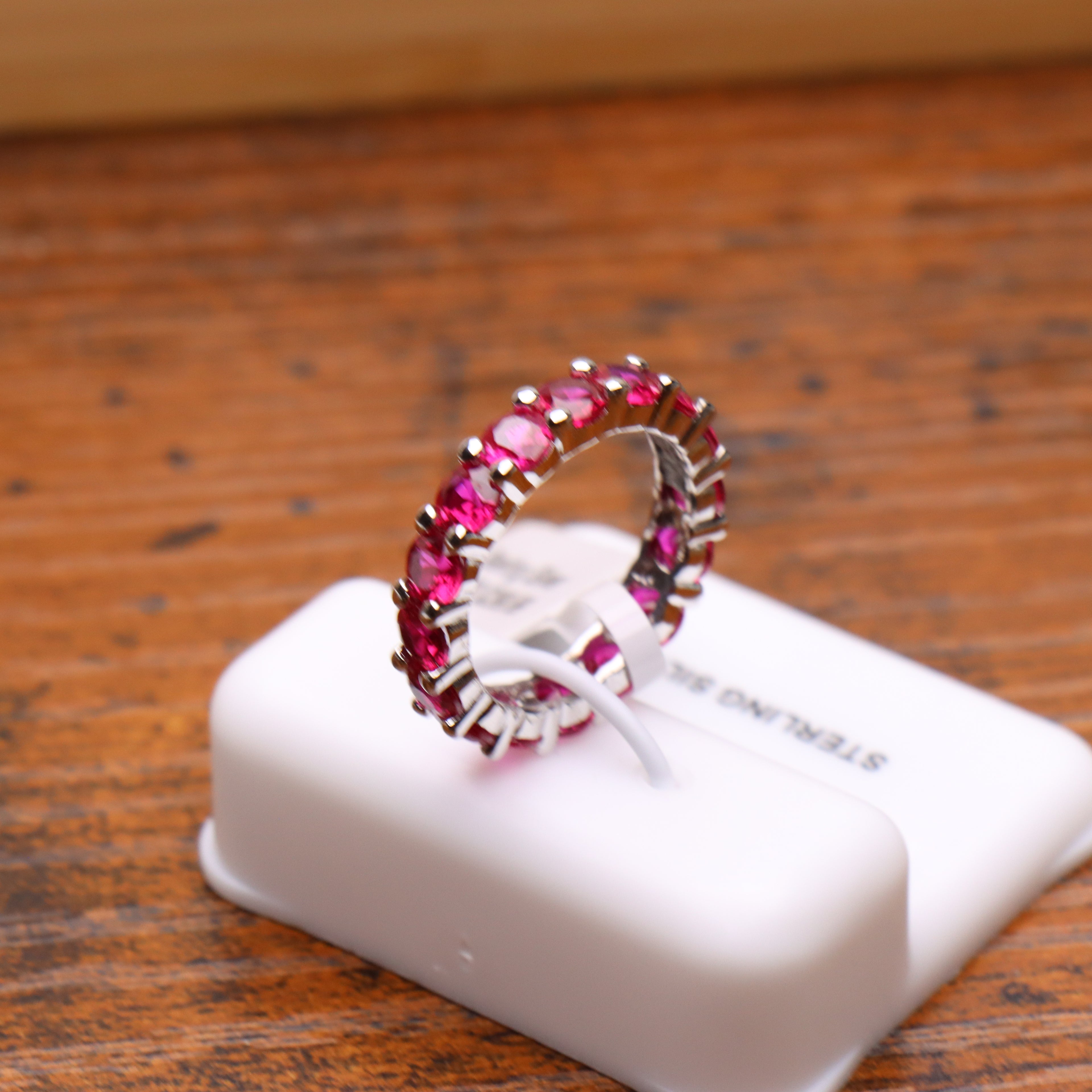 A silver band ring set with a full row of round pink gemstones, displayed on a white jewelry stand on a wooden surface.