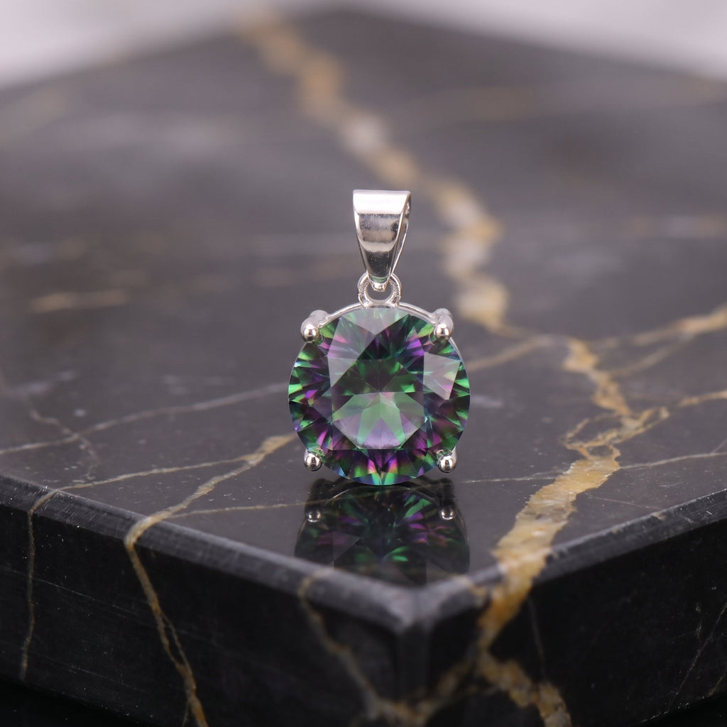 A round-cut iridescent gemstone pendant in a silver four-prong setting, displayed on a polished black marble slab with gold veining. mystic topaz