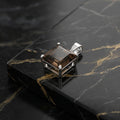 A square smoky brown gemstone pendant in a silver-toned bezel setting, displayed on a black marble slab with gold veining.