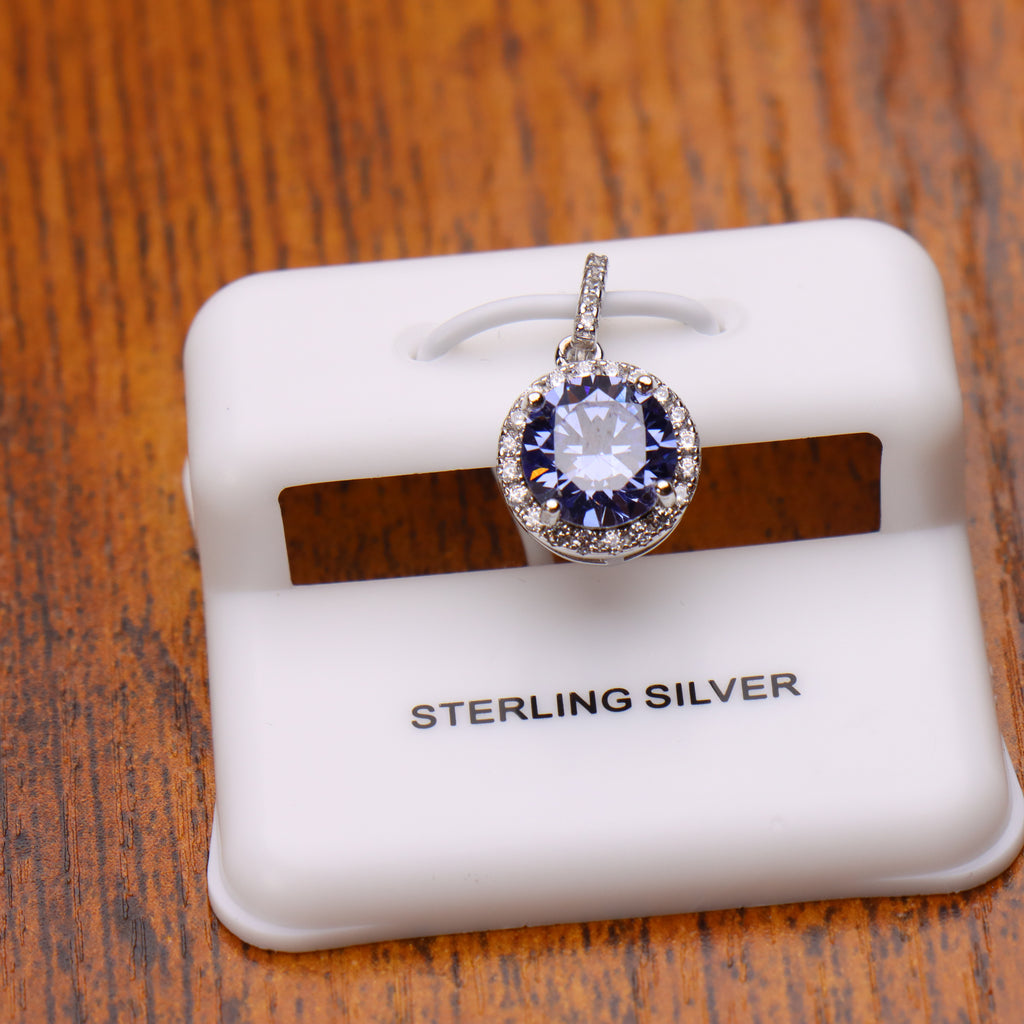 A sterling silver pendant with a round clear central stone surrounded by a halo of small blue stones, displayed on a white jewelry stand over a wooden surface.