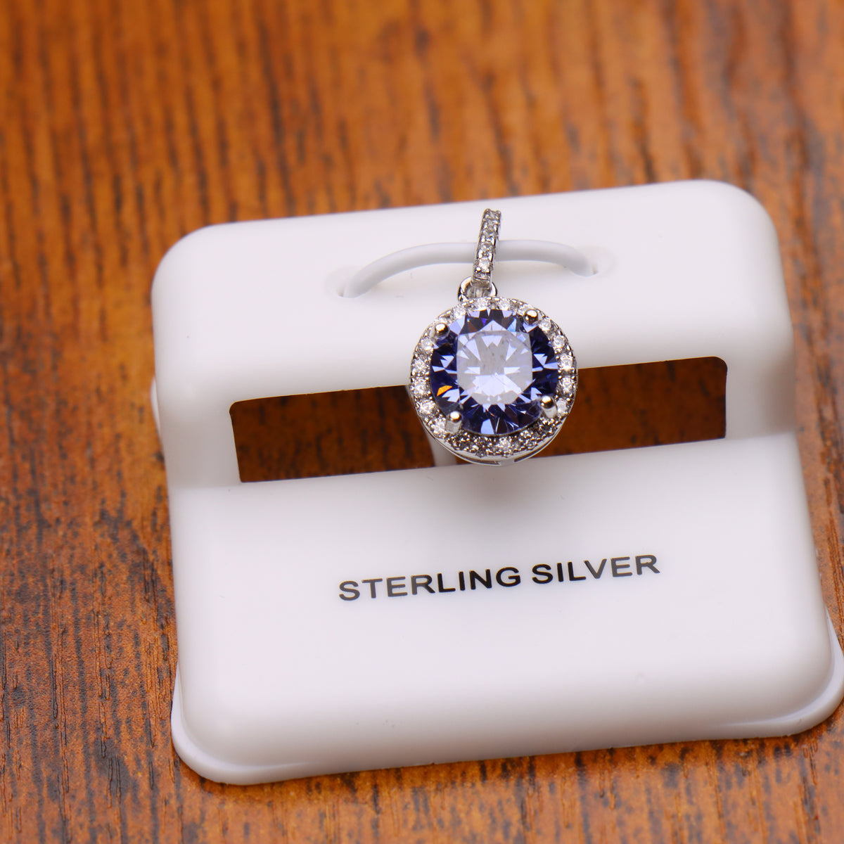 A sterling silver pendant with a round clear central stone surrounded by a halo of small blue stones, displayed on a white jewelry stand over a wooden surface.