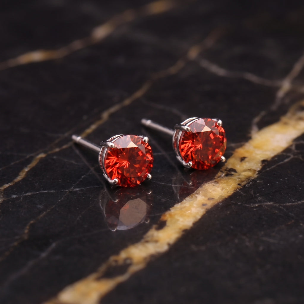 A pair of round-cut red gemstone stud earrings in silver settings, displayed on a polished black marble surface with gold veining.
