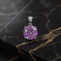 A round-cut purple gemstone pendant in a silver-toned setting, displayed on a black and gold veined marble surface. amethyst