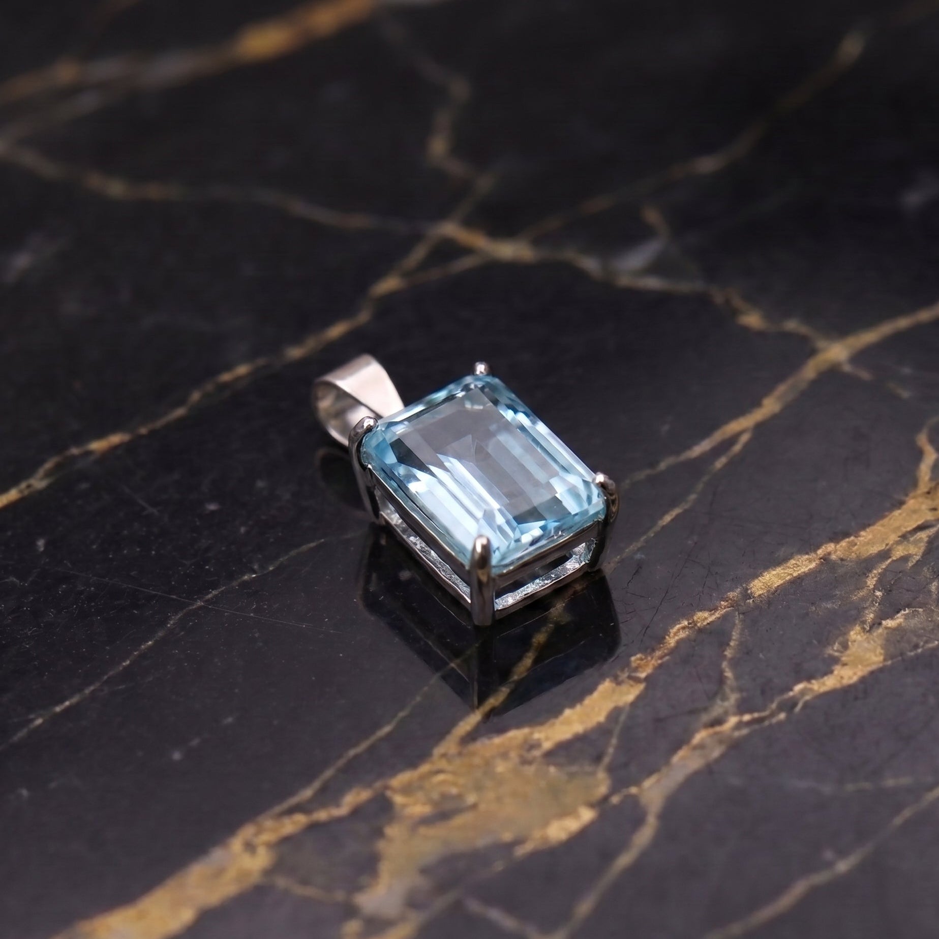A rectangular emerald-cut light blue gemstone pendant in a silver-toned metal setting, displayed on a black marble surface with gold veining.
