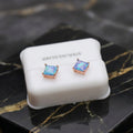 A pair of square stud earrings with iridescent blue-purple stones in rose gold settings, displayed on a white jewelry stand labeled 'STERLING SILVER' on a black marble surface.