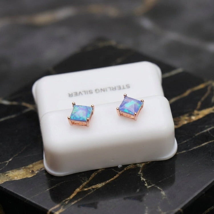 A pair of square stud earrings with iridescent blue-purple stones in rose gold settings, displayed on a white jewelry stand labeled 'STERLING SILVER' on a black marble surface.