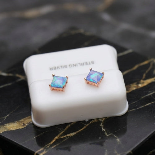 A pair of square stud earrings with iridescent blue-purple stones in rose gold settings, displayed on a white jewelry stand labeled 'STERLING SILVER' on a black marble surface.