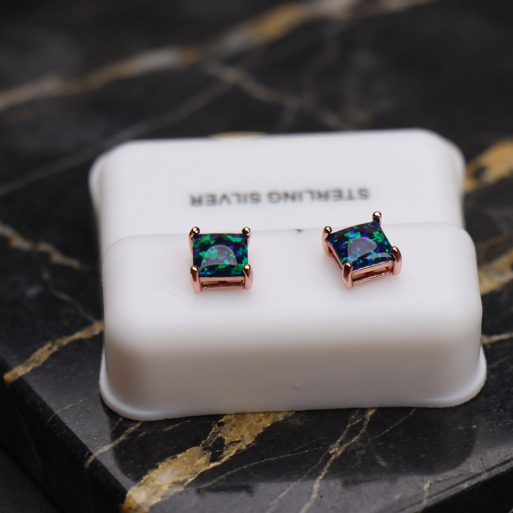 A pair of square stud earrings with iridescent green-blue opal-like centers in rose gold settings, displayed on a white 'STERLING SILVER' box placed on a black marble surface.