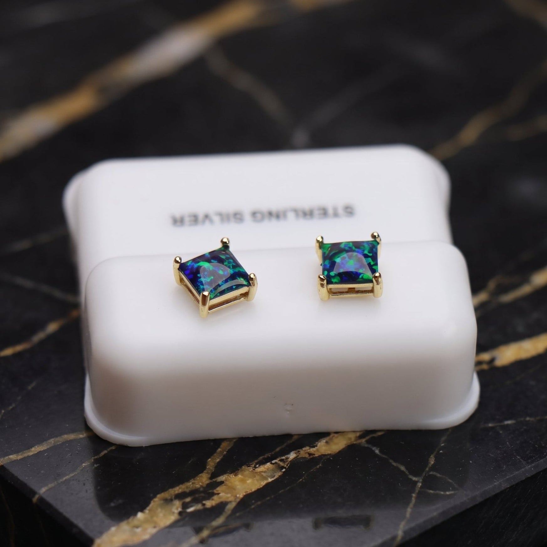 A pair of square stud earrings with iridescent blue-green stones in gold settings, displayed on a white jewelry box over a black marble surface.