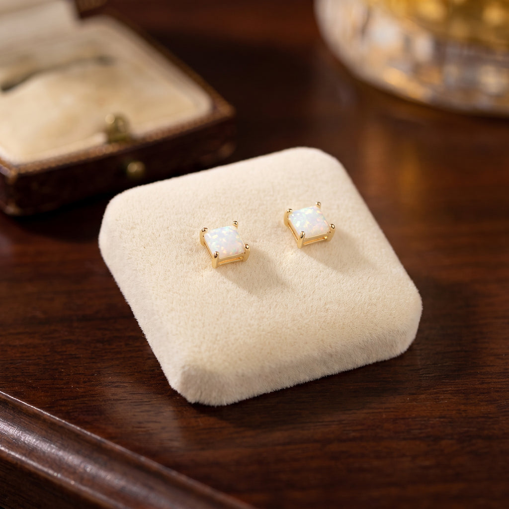 White Opal Square Earrings, featuring lab-created opals set in 14K gold over 925 sterling silver, displayed on a cream-colored velvet pad atop dark wood with a jewelry box and glass object in the background.
