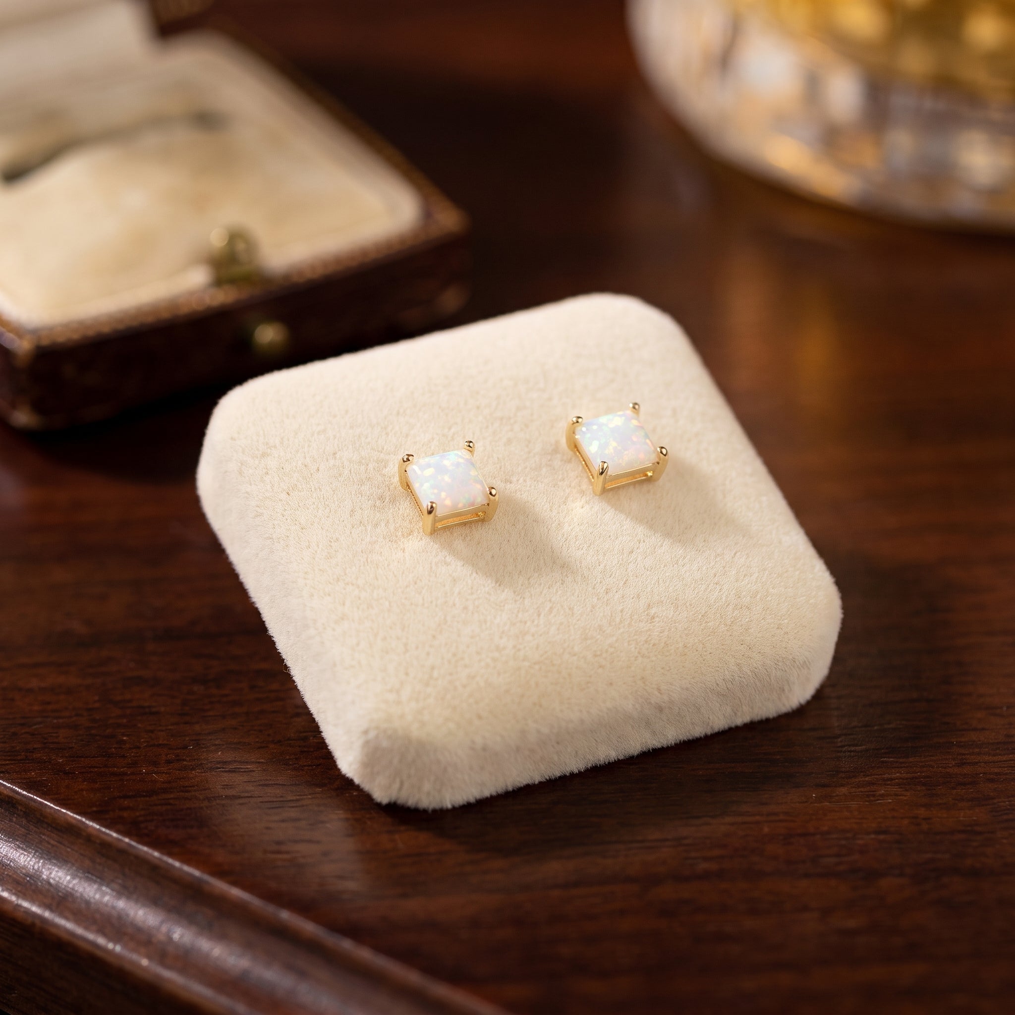 White Opal Square Earrings, featuring lab-created opals set in 14K gold over 925 sterling silver, displayed on a cream-colored velvet pad atop dark wood with a jewelry box and glass object in the background.