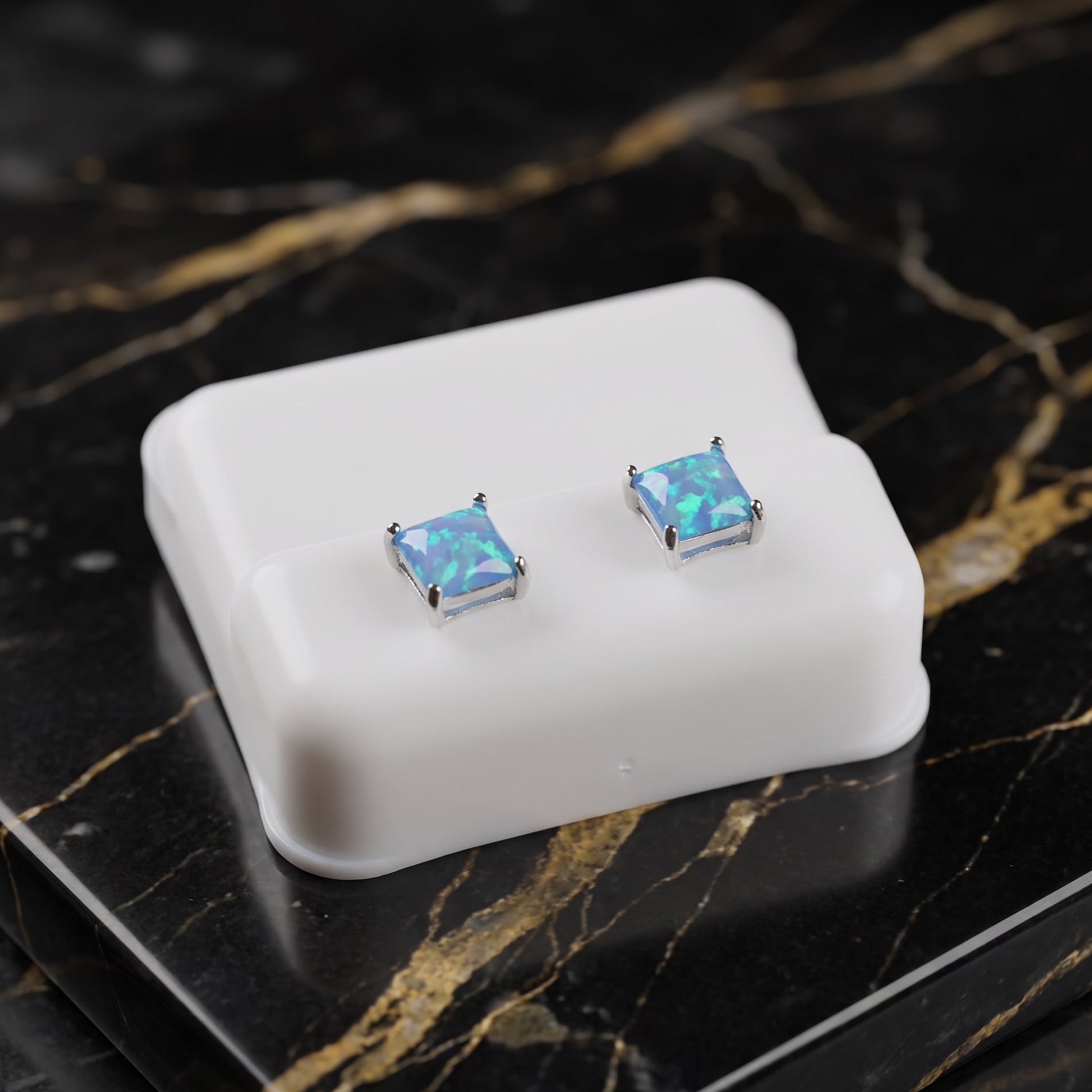 A pair of square stud earrings with iridescent blue opal-like centers in silver settings, displayed on a white rectangular stand over a black marble surface with gold veining.