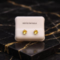 A pair of round yellow gemstone stud earrings in gold settings, displayed on a white square card labeled 'STERLING SILVER' against a black marble background with gold veining.
