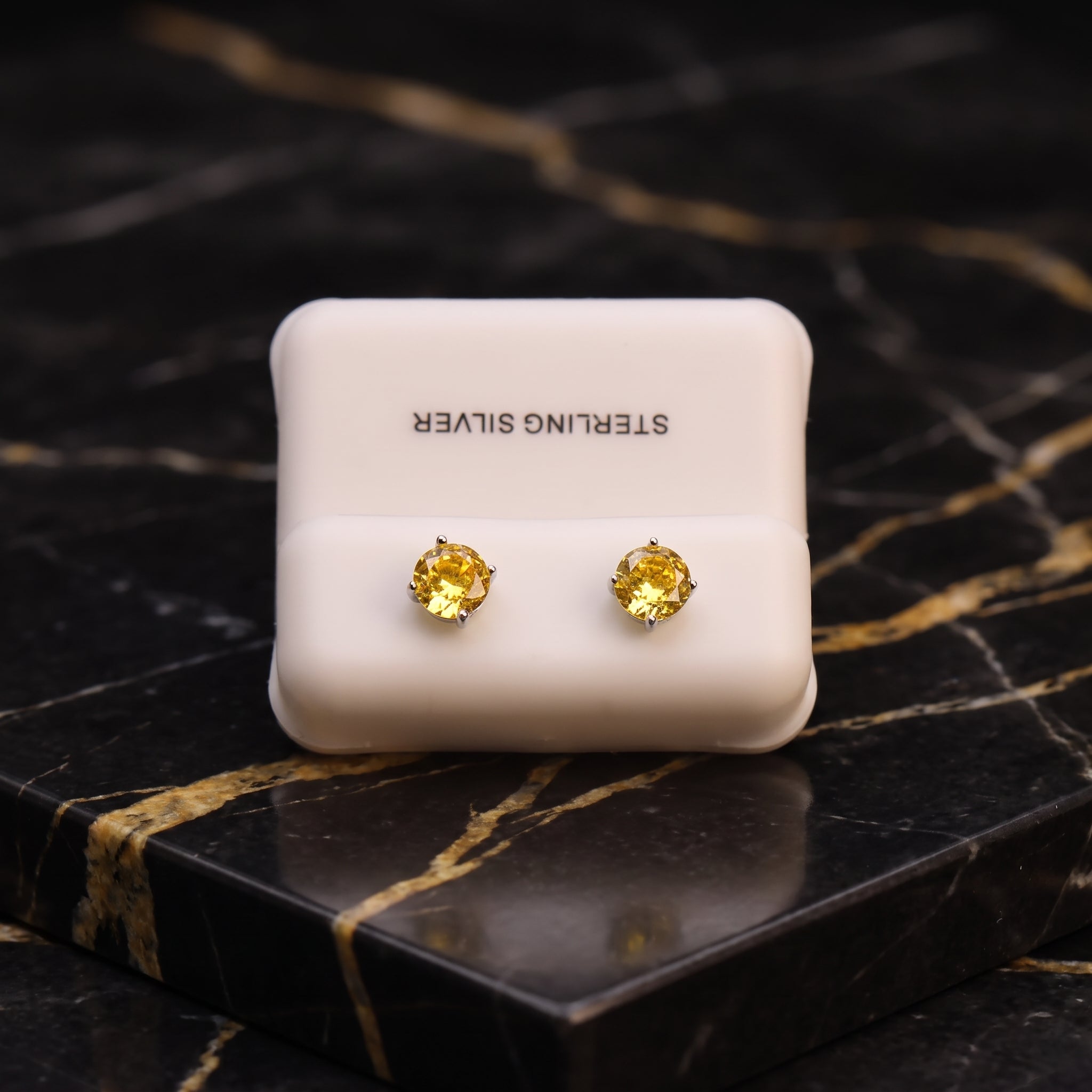 A pair of round yellow gemstone stud earrings in gold settings, displayed on a white square card labeled 'STERLING SILVER' against a black marble background with gold veining.