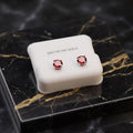 A pair of round-cut red gemstone stud earrings in silver settings, displayed on a white 'STERLING SILVER' branded jewelry box set on a black and gold marble surface.