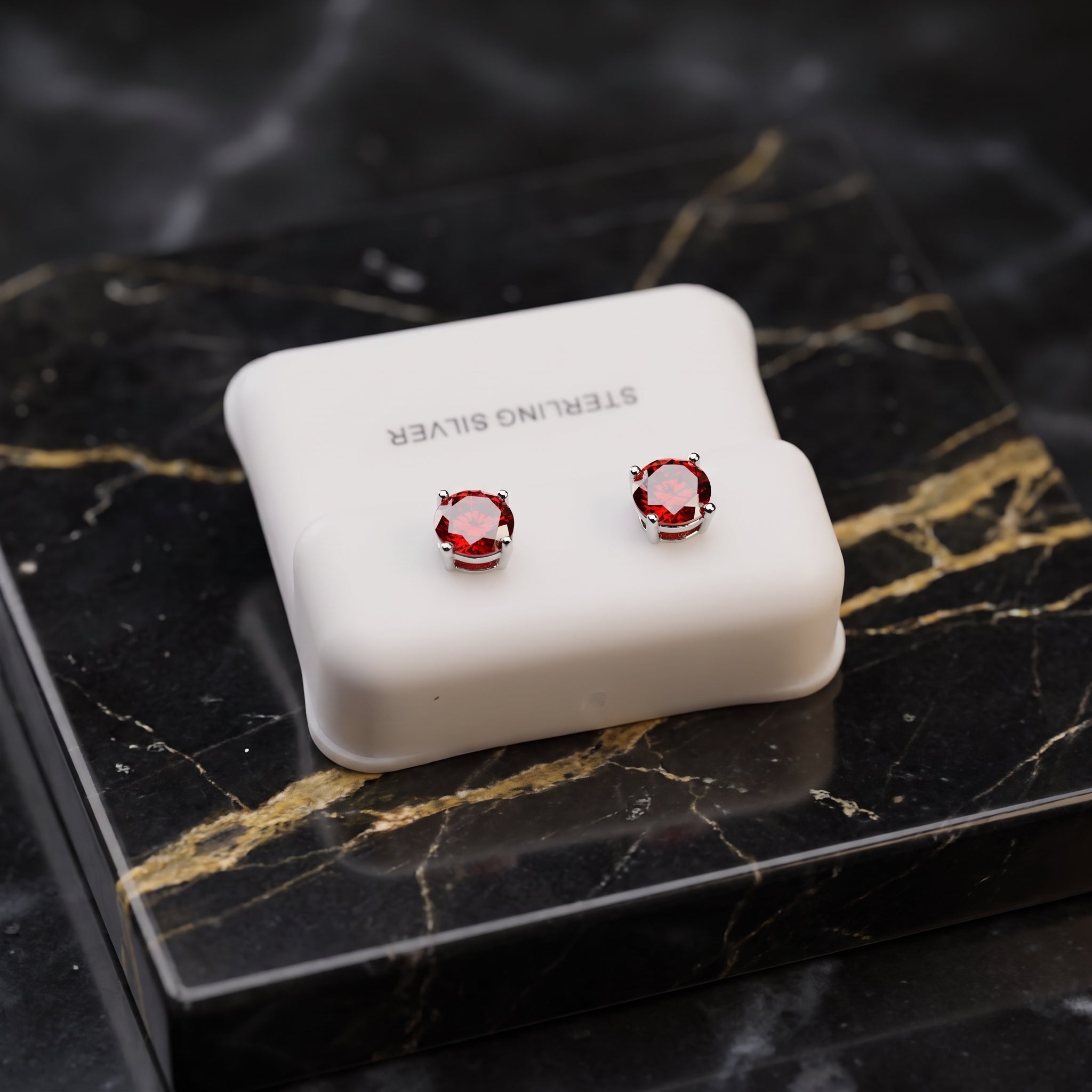 A pair of round-cut red gemstone stud earrings in silver settings, displayed on a white 'STERLING SILVER' branded jewelry box set on a black and gold marble surface.