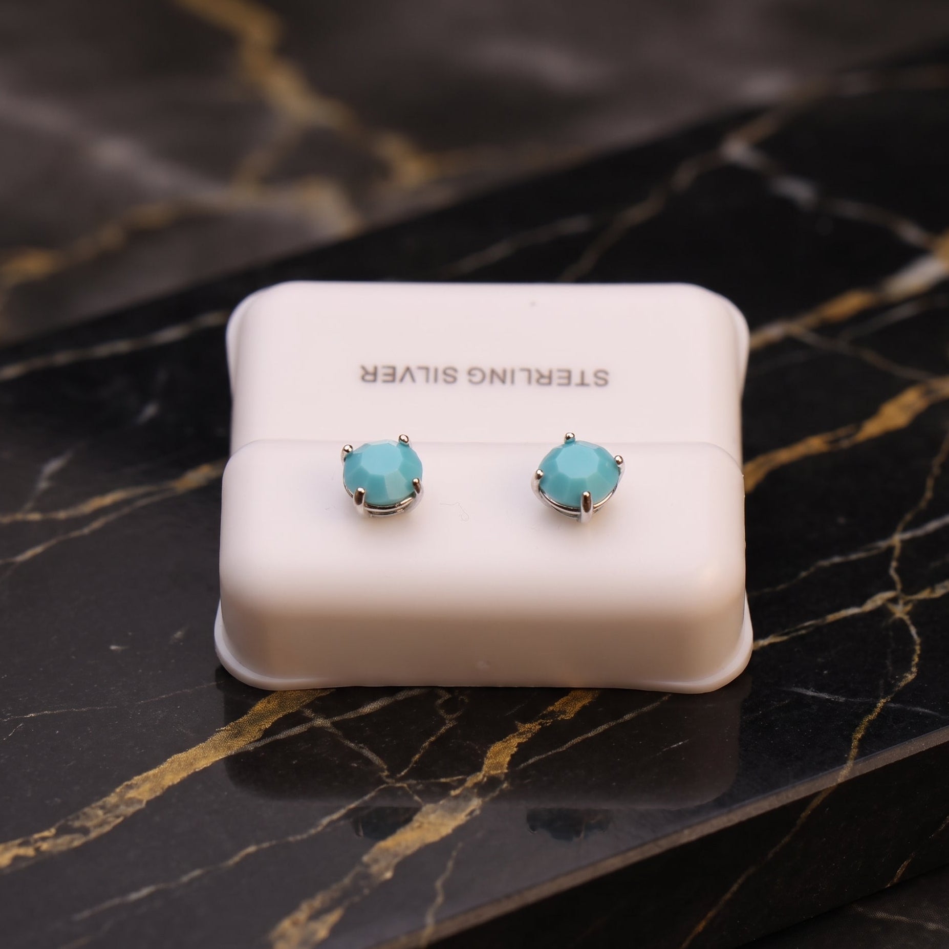 A pair of sterling silver stud earrings with round light blue-green gemstones, displayed on a white jewelry box over a black marble surface with gold veining.